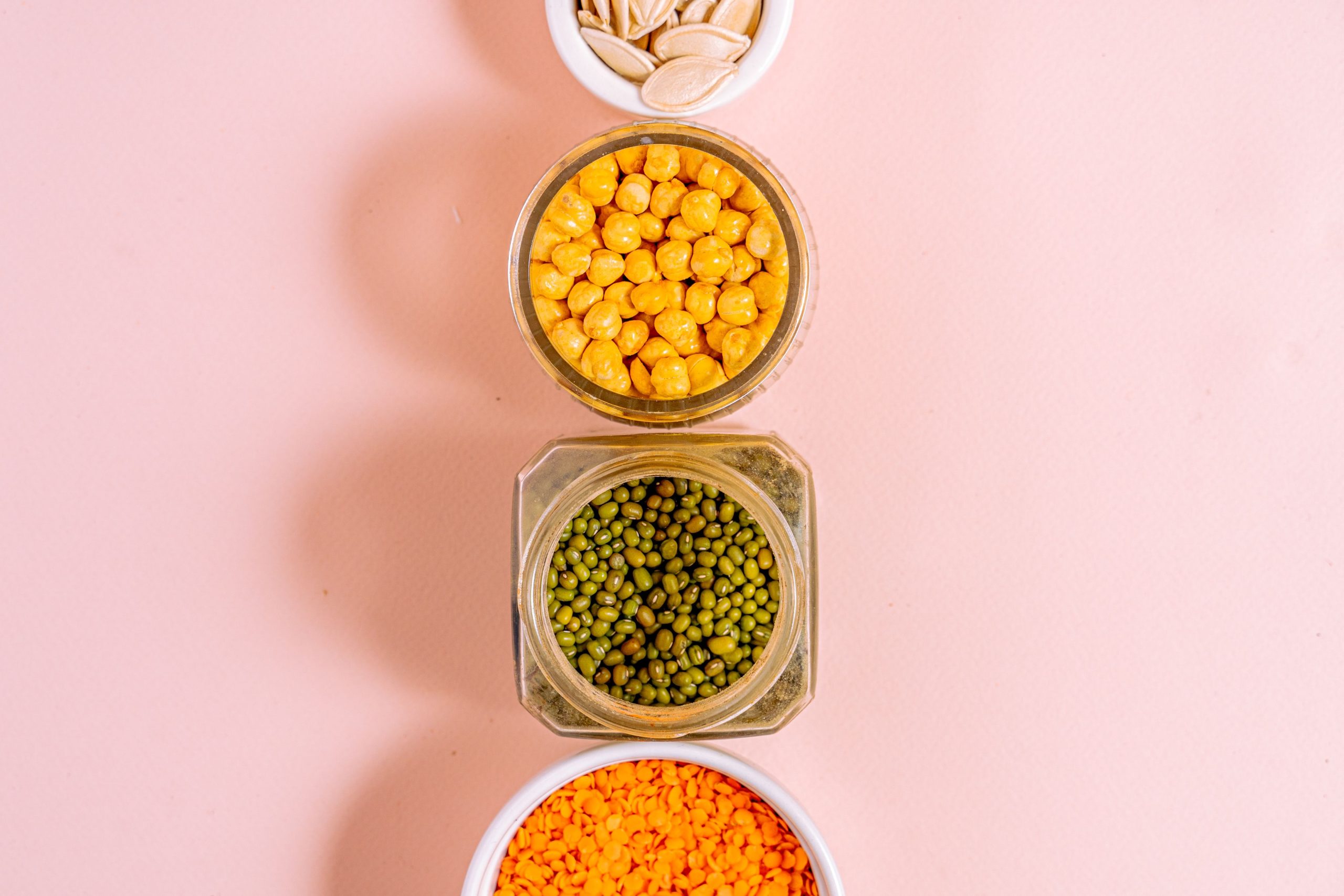 Top view of assorted legumes and seeds in jars on a pink background, emphasizing healthy eating.