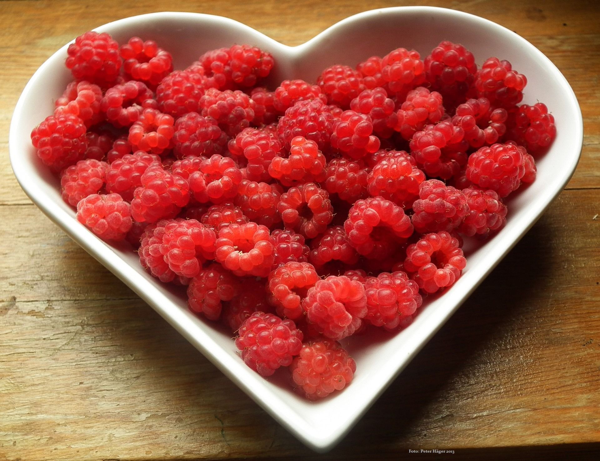 Vibrant red raspberries in a heart-shaped bowl on a wooden surface, perfect for food and health themes.